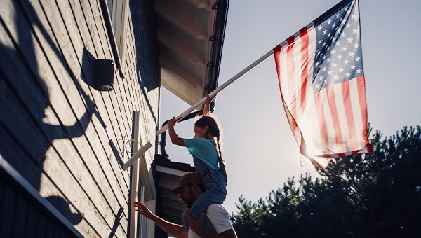 A daughter sits on the shoulders of her father to place an American flag into a holder on the side of a house.