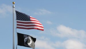 An American flag and POW/MIA flag fly in front of a blue sky and clouds.