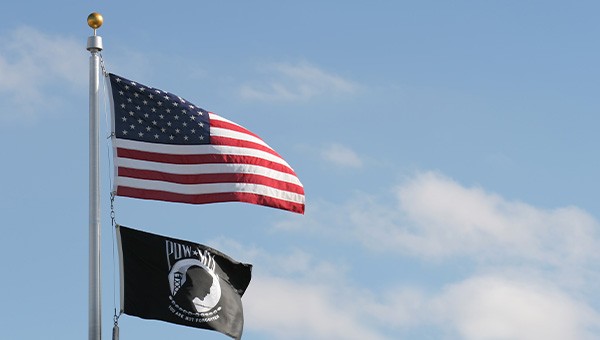An American flag and POW/MIA flag fly in front of a blue sky and clouds.