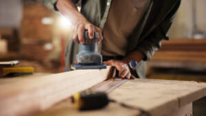 Person, hands and carpenter with sander at workshop for polish, craft or lumber project. Closeup, wood worker and sanding machine with plank, timber or beam for wooden repair, design or restoration.