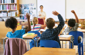 Education, question with group of children in classroom and raise their hands to answer. Learning or support, diversity and teacher teaching with young students in class of school building together.